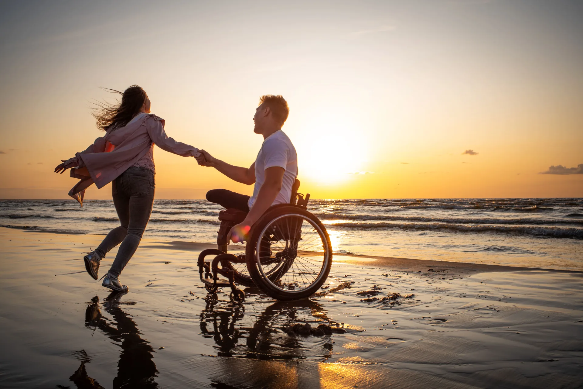 A man in a wheelchair and a woman holding hands on the beach at sunset