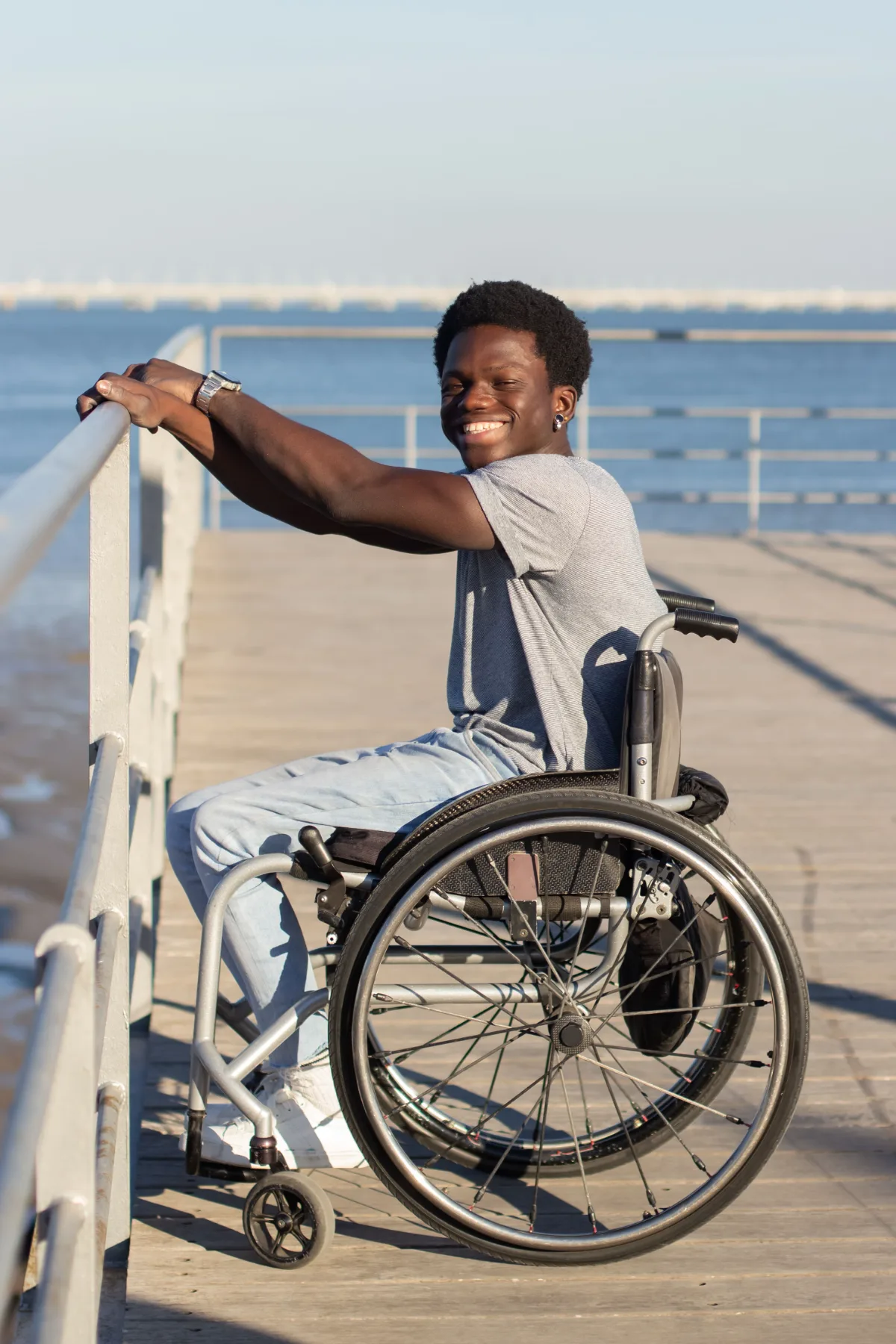 A young man in a wheelchair smiling on a boardwalk by the water