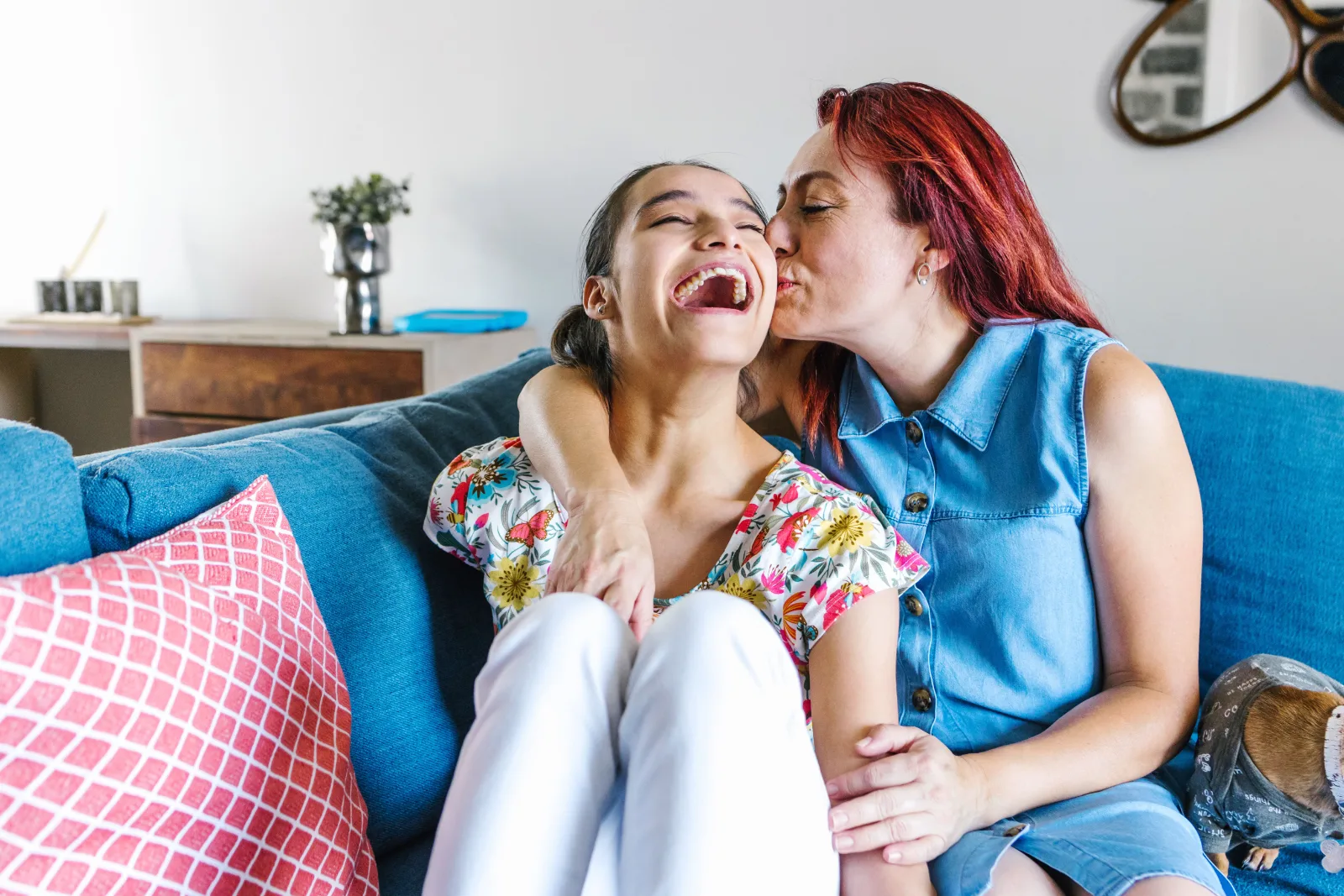 A mother kissing her laughing teenage daughter on the cheek at home