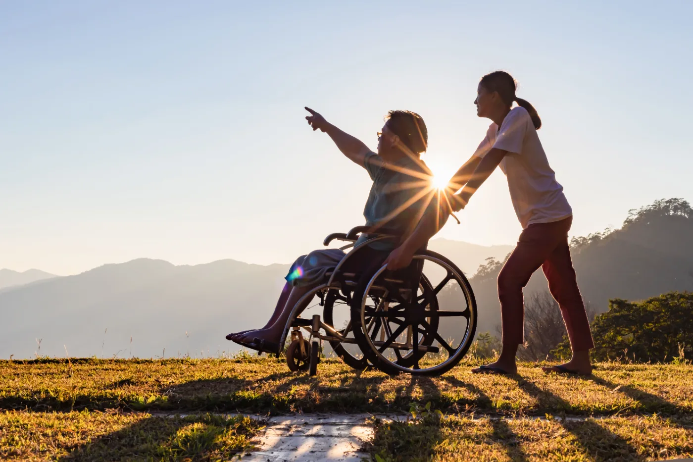 A support worker and a man in a wheelchair looking out over hills at sunset